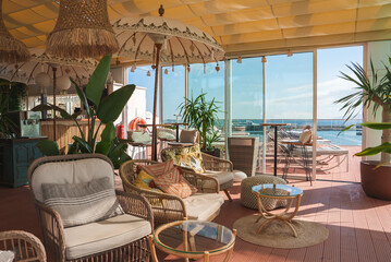 Indoor lounge area features wicker furniture with colorful cushions, decorative umbrellas, and lush plants. Glass windows offer a view of the pool area.