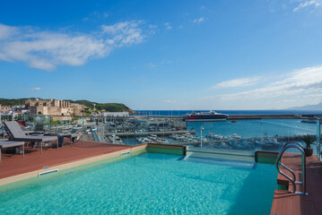 Fototapeta premium A rooftop pool area with lounge chairs overlooks a marina, featuring yachts and a ferry. A historic fortress and distant hills are visible in the background.
