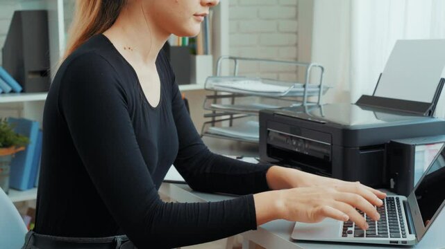 Woman using printer to print documents in modern home office with laptop on desk and organized workspace