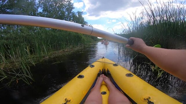 Person floating in inflatable boat along river through thickets of green reeds on sunny summer day. People kayaking along river. Man in boat rowing oars. Sport, action, activity, lifestyle. POV