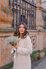 Beautiful, elegant, stylish woman in silk dress and fur coat with white bouquet walking in the city 