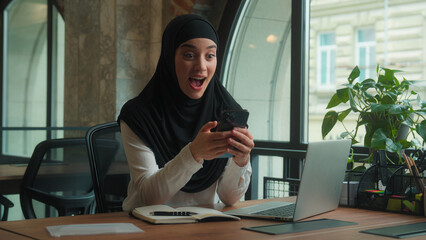 Arabian muslim islamic woman in hijab female businesswoman happy business girl employer winner excited amazed using mobile phone good news achieve yes win smartphone online victory success in office