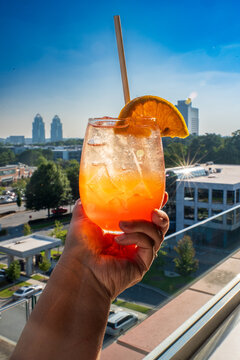 A hand is holding a glass of coctail. A vibrant mix of colors, with layers of deep red, bright orange, topped with a slice of citrus fruit. Background of iconic Queen, King buildings, downtown Atlanta