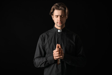 Young priest praying with beads on dark background