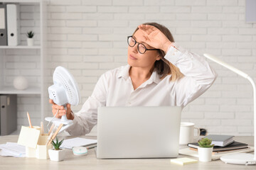 Beautiful mature businesswoman with electric fan and laptop suffering from heat in office