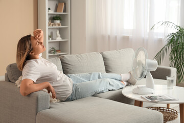 Fototapeta premium Beautiful mature woman with electric fan suffering from heat on sofa at home