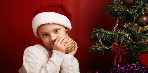 Smiling cute kid girl holding in hands Christmas tree ornament golden ball near the holiday fur green tree in santa claus hat. Happy expression winter holidays. Closeup portrait