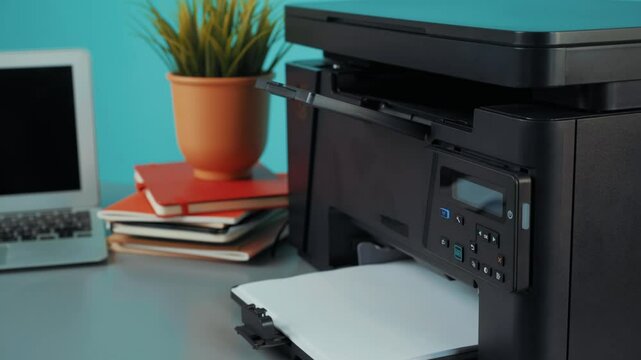 Home office setup with multifunction printer and laptop on desk with potted plant and stacked notebooks for productivity