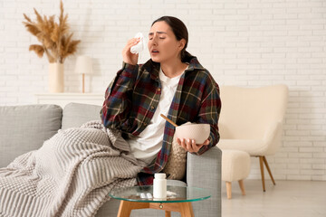 Sick young woman with tissue and soup sneezing on sofa at home