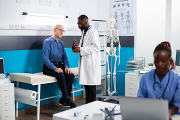 Obraz premium Black doctor offers health advice to senior white man during medical checkup holding bottle of prescribed pills. Pensioner patient receives friendly consultation on healthcare plan and medication.