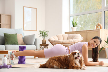 Sporty young woman exercising with cute cavalier King Charles spaniel on yoga mat at home