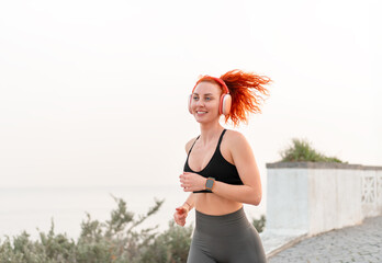 Happy sporty jogger woman jogging against seascape against clear sky. Smiling redheaded female in...