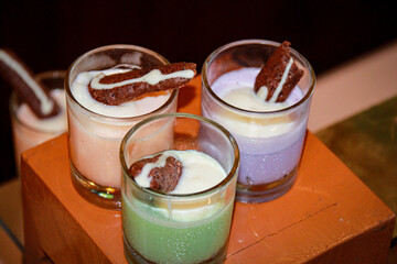 Assorted puddings in green, white, and pink with chocolate biscuits as garnish, served in glass...