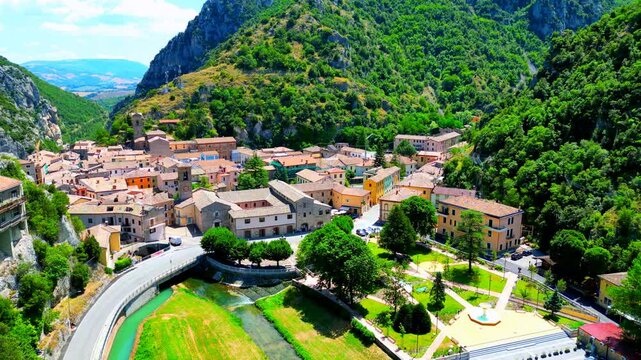 Stunning aerial pedestal up shot from Pioraco encompassing the Potenza river passing under bridges, green park with trees, road with cars, town center with close buildings and huge Sibillini mountains