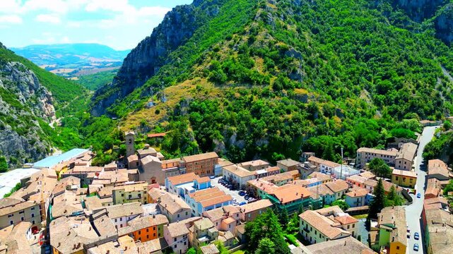 Breezy aerial pan left push-in shot from Pioraco, a fly-over vision of the town center with tight buildings, roads with passing cars, technological towers, factories, great Sibillini mountains' gorge