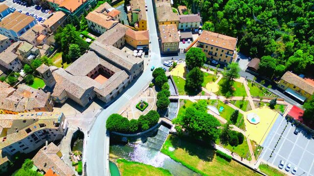 Geometrical aerial top-down bird's-eye view of the Pioraco town centre, a moving vision of the park, fountain, trees, the Potenza river, town hall, cloister, belltower, thriving vegetation, road, cars