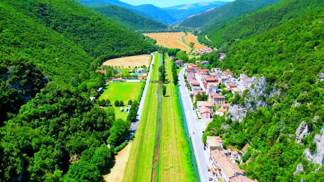 Vast aerial tilt-up push-in shot from Pioraco, swathes of green shores flanking the Potenza river, fields, buildings, and the impressive, far-reaching Sibillini mountains all around under puffy clouds