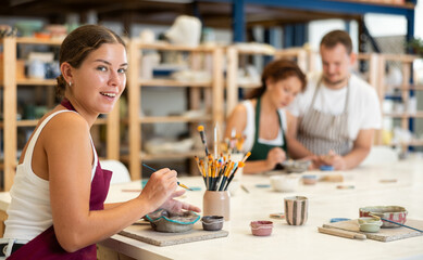 Concentrated girl in apron designing handmade clay plate with brush in taller room