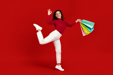 A young Caucasian woman joyfully poses against a bright red background, holding colorful shopping...