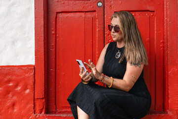 Stylish woman with tattoos using smartphone in front of a red rustic door outdoors