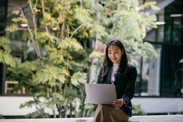 Young asian businesswoman smiling and using laptop while sitting in modern office space with green plants, enjoying work-life balance in natural environment