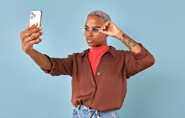 Confident African American individual poses against pastel blue backdrop, taking a stylish selfie. With vibrant attire and playful expression, they showcase their unique fashion sense and personality