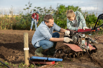 Male farmer shows his son how to fix garden cultivator. Farmer and a guy are repairing a machine to...