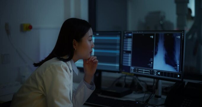 Professional Radiologist Studies Patient X-rays in an Advanced Software on a Computer Display in a Darkened Room. Asian Medical Specialist Making an Assessment of a Critical Medical Data