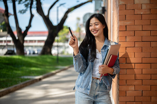 A female student stands leaning against a brick building wall, smiling and looking to the side. - Powered by Adobe