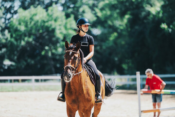 A young equestrian wearing a helmet riding a horse in an outdoor arena on a sunny day. The setting is peaceful and suitable for equestrian training and leisure activities.