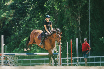 A young equestrian skillfully guides a horse over a jumping hurdle in an outdoor arena,...