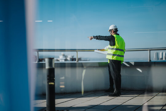 Two architects wearing safety gear discuss building plans while pointing at a construction site. The rooftop view emphasizes city planning and development. The scene conveys professionalism and