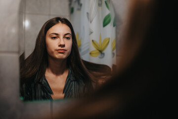 A young woman gazes thoughtfully at her reflection in the bathroom mirror. Her expression conveys...