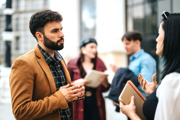 A diverse group of business people engaged in a serious outdoor conversation. They hold notebooks,...