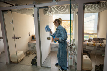 Female worker diligently cleans glass doors in cat rooms with sprayer, ensuring dust free, pleasant setting for animals in hotel. High quality lodging for cats, combining hygiene with pet comfort