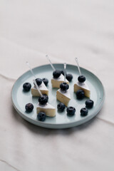 In the image, a gloved hand is delicately placing a blueberry on a piece of cheese. 