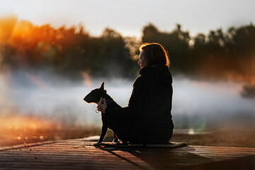 miniature bull terrier sitting with owner by lake in early foggy sunrise