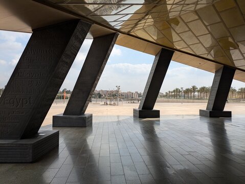 New Grand Egyptian Museum stark schr&auml;g stehenden, massiven St&uuml;tzen zu sehen, der golden schimmernde Eingang zum Museum, Palmen und vereinzelten Strukturen in der Ferne unter leicht bew&ouml;lktem Himmel.