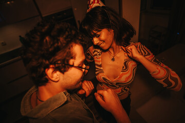 A young couple joyfully dances together in a warmly lit home setting, celebrating a birthday. Both are smiling, wearing festive attire, and enjoying an intimate and fun moment.