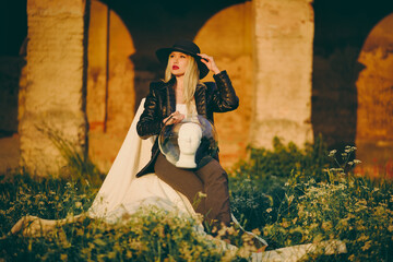 Woman blonde in hat sitting on chair in nature on background of old building with arches. Lady is holding an aquarium and a mannequin head in her hands. Fashion concept glamor.