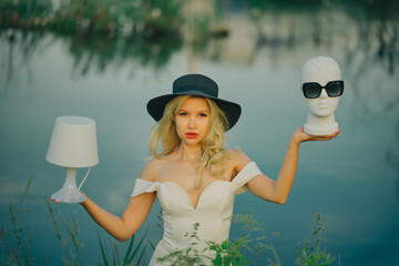 Portrait of a beautiful woman in white dress and black hat near a pond. Glamorous lady holding floor lamp and mannequin head with glasses.