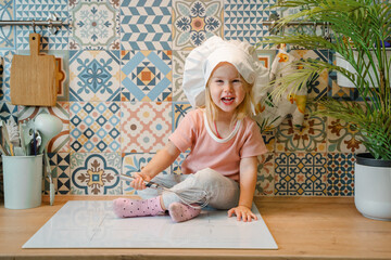 A blonde Caucasian child girl toddler with a whisk in the kitchen sits on the countertop and helps...