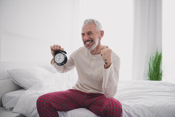 Joyful mature man in pajamas sitting on bed with alarm clock embracing morning light and relaxation