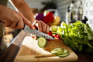 Close-up of hands slicing cucumber with fresh vegetables on cutting board