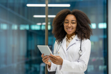 A young woman doctor stands confidently in a modern medical office, using a tablet to access...
