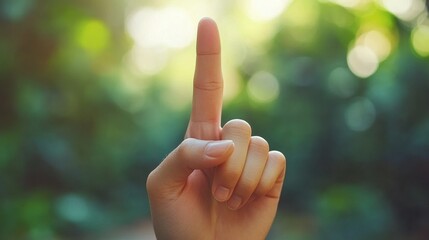 A hand is shown with one finger raised, signaling or emphasizing something important amid a natural, blurred green backdrop under sunlight