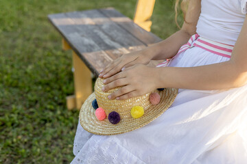 Girl in white dress sits on wooden bench laying her straw hat on knee, summer outdoor lifestyle