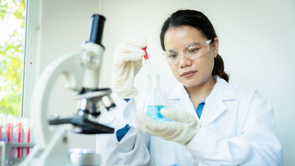 Fototapeta premium A woman in a lab coat is holding a test tube with a blue liquid in it. She is wearing gloves and a face mask, and is looking at the tube with a serious expression