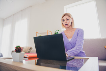 Fototapeta premium Young woman wearing a knitted sweater looking surprised at laptop in bright living room during daytime