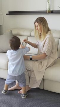 young blonde woman mother sits on sofa uses laptop. little kid son comes and closes laptop. protests does not let work. raising children theme. video footage